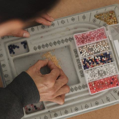 A close-up overhead view of a person's hands working with small colorful beads inside a gray segmented tray. A clear plastic organizer box filled with pink, white, blue, and multicolored beads sits to the right.