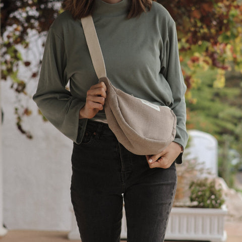 Person holding a beige crossbody bag outdoors with greenery in the background