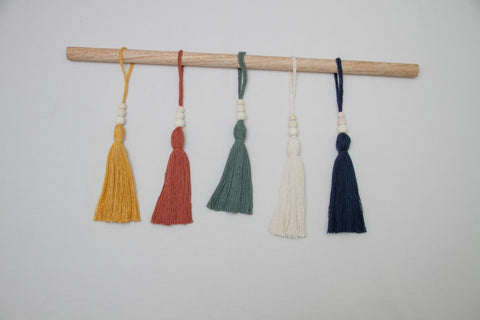 Not I But We's small tassel ornaments in mustard, green, navy, terra, and cream presented on a stick.