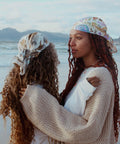 Two models wearing Not I But We's satin scarf around their heads at the beach in Muizenberg.