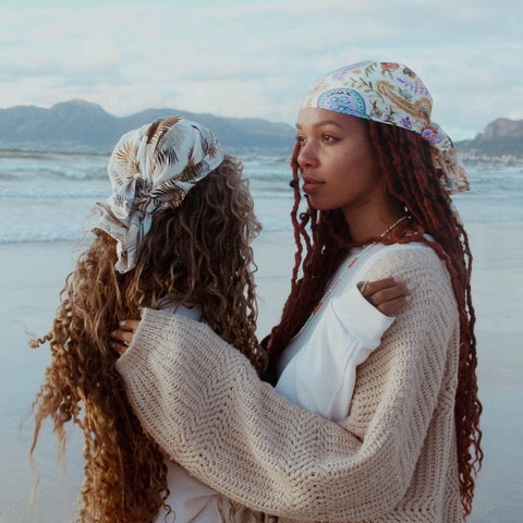 Two models wearing Not I But We's satin scarf around their heads at the beach in Muizenberg.