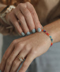 A close-up lifestyle shot of a person's hands and wrists, showcasing a colorful beaded bracelet on the wrist along with gold rings.