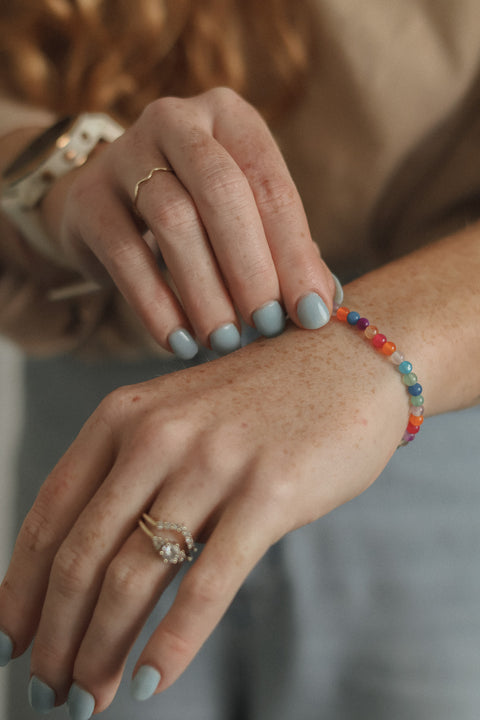 A close-up lifestyle shot of a person's hands and wrists, showcasing a colorful beaded bracelet on the wrist along with gold rings.