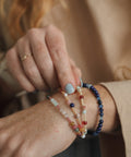 A close-up of a person's wrist wearing three delicate beaded bracelets, including a navy blue bead bracelet, a light blue and white beaded bracelet, and a peach and red beaded bracelet, against a soft beige background.