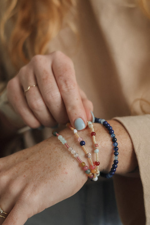 A close-up of a person's wrist wearing three delicate beaded bracelets, including a navy blue bead bracelet, a light blue and white beaded bracelet, and a peach and red beaded bracelet, against a soft beige background.