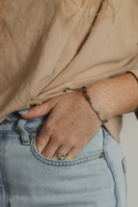 A close-up lifestyle shot of a person wearing a delicate beaded bracelet on their wrist, paired with a beige top and light blue jeans.