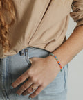 A close-up lifestyle shot of a person wearing a thin beaded bracelet with multicolor beads on their left wrist, paired with a beige top and light blue jeans.