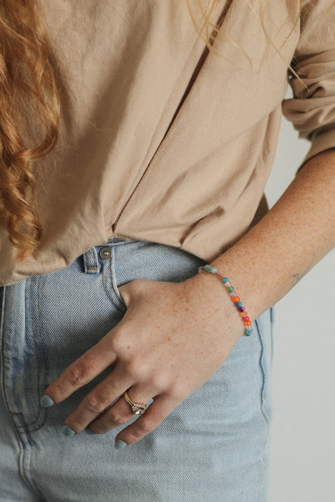 A close-up lifestyle shot of a person wearing a thin beaded bracelet with multicolor beads on their left wrist, paired with a beige top and light blue jeans.