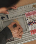 A close-up overhead view of a person's hands working with small colorful beads inside a gray segmented tray. A clear plastic organizer box filled with pink, white, blue, and multicolored beads sits to the right.