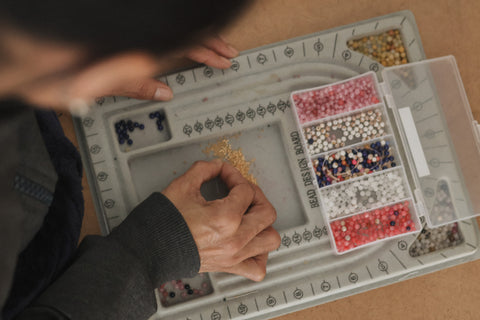 A close-up overhead view of a person's hands working with small colorful beads inside a gray segmented tray. A clear plastic organizer box filled with pink, white, blue, and multicolored beads sits to the right.