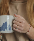 A close-up lifestyle shot of a person holding a white ceramic mug with a blue abstract brushstroke pattern. The person is wearing a beige top, a gold ring, and a beaded bracelet.
