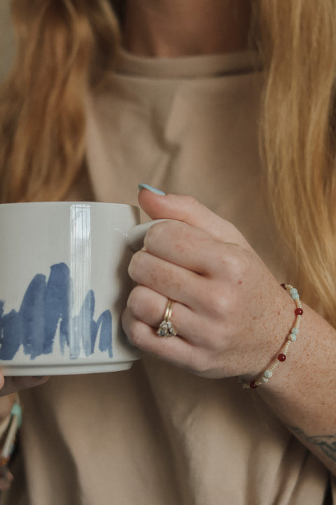 A close-up lifestyle shot of a person holding a white ceramic mug with a blue abstract brushstroke pattern. The person is wearing a beige top, a gold ring, and a beaded bracelet.