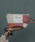 A hand wearing a beige knit sleeve and bracelets holds a small patchwork zip pouch against a textured dark teal wall. 