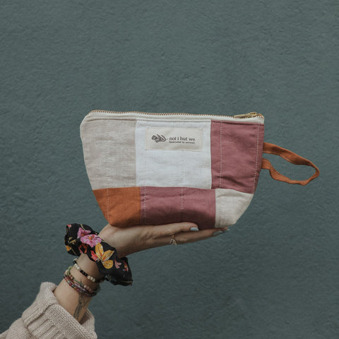 A hand wearing a beige knit sleeve and bracelets holds a small patchwork zip pouch against a textured dark teal wall. 